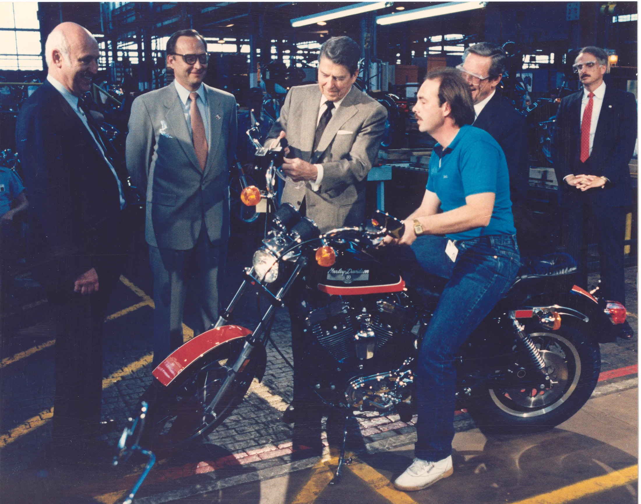Color photo taken during President Reagan's May 1987 visit to York. Ronald Reagan with Vaughn Beals and Richard Teerlink, all standing next to a motorcycle smiling, in the plant, with factory worker.